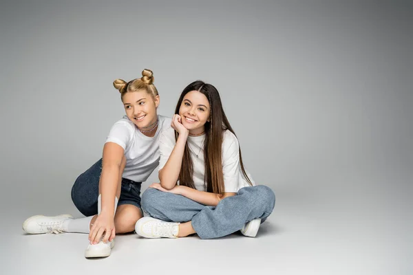 Cheerful and teen girlfriends in white t-shirts, sneakers and denim shorts smiling at camera while sitting together and posing on grey background, lively teenage girls concept, friendship and bonding — Stock Photo
