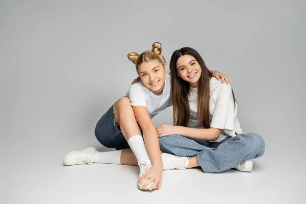 Smiling blonde teenage girl in white t-shirt and denim shorts hugging girlfriend and looking at camera while sitting on grey background, lively teenage girls concept, friendship and bonding — Stock Photo