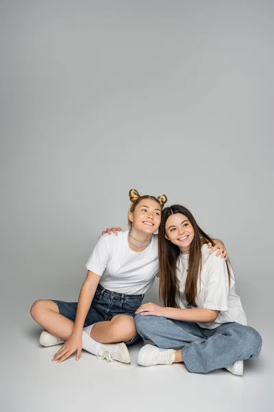 Радостные и подростковые девушки в белых футболках и кроссовках, обнимающие друг друга, отводящие взгляд и сидящие вместе на сером фоне, живая концепция девочек-подростков, дружба и дружба — стоковое фото