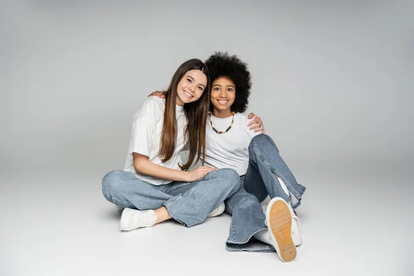 Lächelndes brünettes Teenie-Mädchen in weißem T-Shirt und Jeans umarmt afrikanisch-amerikanische Freundin, während sie zusammen sitzt und in die Kamera auf grauem Hintergrund schaut, lebhaftes Teenager-Mädchen-Konzept — Stockfoto
