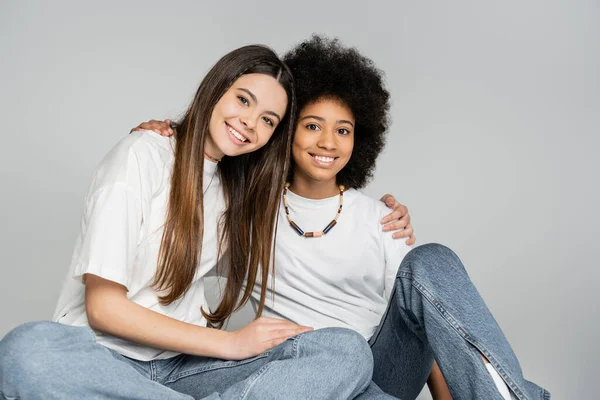 Радостная девушка-подросток в обычной белой футболке и джинсах, обнимающая африканскую американскую подружку и смотрящая в камеру, позируя изолированной на серой, живой концепции девочек-подростков, дружбе и связи — стоковое фото