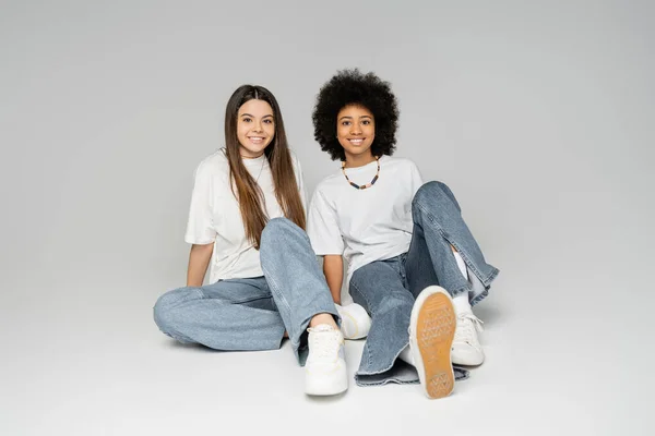 Full length of positive african american teenage girl in white t-shirt and blue jeans sitting next to brunette girlfriend on grey background, lively teenage girls concept, friendship and bonding — Stock Photo