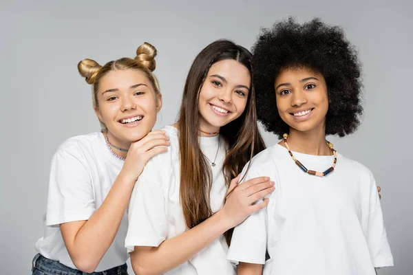 Porträt fröhlicher, stilvoller und multiethnischer Teenager-Freundinnen in weißen T-Shirts, die gemeinsam in die Kamera schauen, während sie isoliert auf grauen, lebhaften Teenagermädchen stehen. — Stockfoto