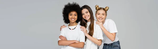 Positive multiethnic teen girlfriends in casual t-shirts and jeans hugging each other and looking at camera together isolated on grey, lively teenage girls concept, banner with copy space — Stock Photo