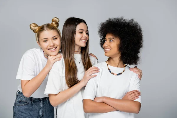 Блондинка и брюнетка девушки-подростки в белых футболках, обнимающие уверенную африканскую американскую подружку, скрещивающую руки изолированные на серой, живой концепции девочек-подростков, дружбе и связи — стоковое фото