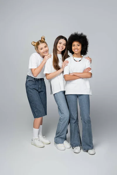 Full length of positive and interracial teenagers in casual white t-shirts and jeans looking at camera and standing on grey background, lively teenage girls concept, friendship and bonding — Stock Photo