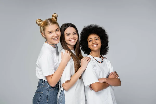 Fröhliche blonde und brünette Teenager in weißen T-Shirts umarmen ihre afrikanisch-amerikanische Freundin, während sie isoliert auf grauen, lebhaften Teenagermädchen stehen. — Stockfoto