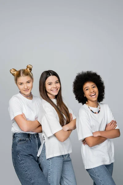Confident and cheerful multiethnic girlfriends in casual white t-shirts and jeans crossing arms and looking at camera while standing isolated on grey, lively teenage girls concept — Stock Photo
