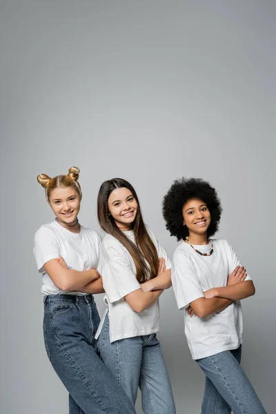 Позитивні і багатоетнічні дівчата-підлітки в стильних білих футболках і джинсах дивляться на камеру і схрещують руки, стоячи разом ізольовані на сірій, жвавій концепції дівчаток-підлітків — стокове фото