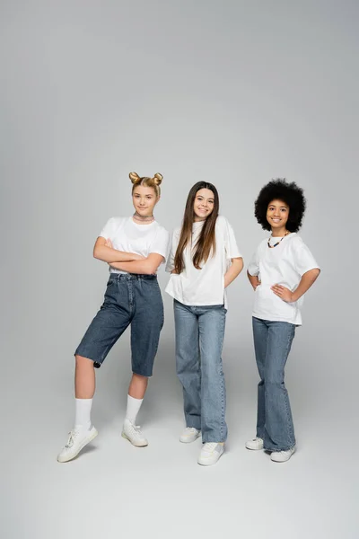 Full length of cheerful and multiethnic teenage girlfriends in white t-shirts and jeans posing while standing together on grey background, lively teenage girls concept, friendship and bonding — Stock Photo