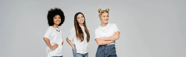 Радостные и многоэтнические девушки-подростки в белых футболках позируют и смотрят в камеру, стоя рядом друг с другом изолированно на серой, живой концепции девочек-подростков, баннер — стоковое фото