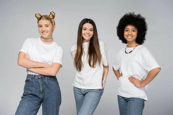 Positive und multiethnische Teenager-Mädchen in Jeans und lässigen weißen T-Shirts posieren und lächeln in die Kamera, während sie isoliert auf grauen, lebhaften Teenagermädchen stehen. — Stockfoto
