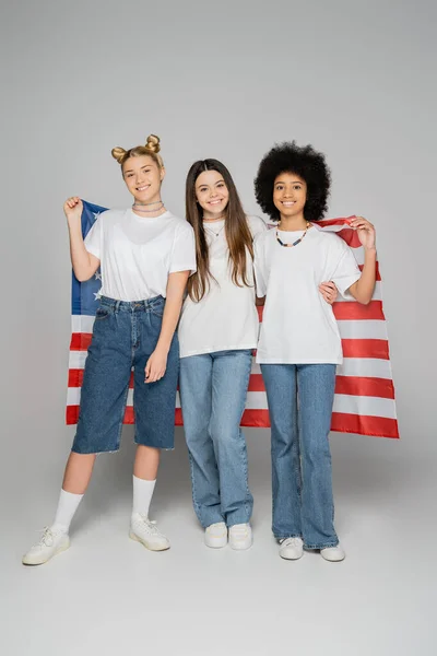 Full length of multiethnic teen girlfriends in white t-shirts and jeans holding ameican flag and looking at camera on grey background, lively teenage girls concept, friendship and companionship — Stock Photo