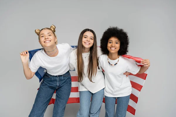 Joyful and interracial teenage girls in white t-shirts and jeans holding american flag and looking at camera while standing on grey background, lively teenage girls concept, friendship — Stock Photo