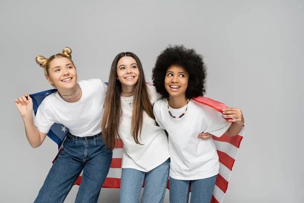 Adolescenti e ragazze multietniche in magliette bianche e jeans sorridenti mentre tengono la bandiera americana e in piedi su sfondo grigio, energici amici adolescenti che trascorrono tempo, amicizia — Foto stock