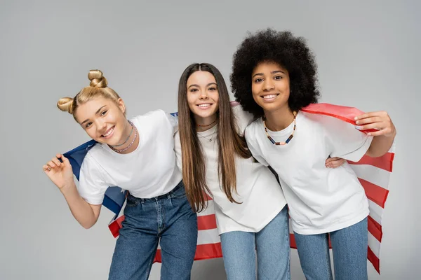 Positive multiethnische Teenager-Freundinnen in weißen T-Shirts und Jeans, die in die Kamera schauen, während sie mit amerikanischer Flagge auf grauem Hintergrund posieren, energische Teenager-Freundinnen, die Zeit verbringen, Freundschaft — Stockfoto