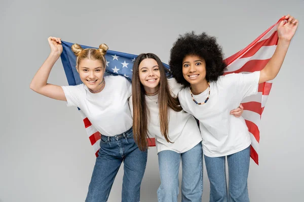 Joyful and multiethnic teenage girlfriends in casual white t-shirts and jeans looking at camera while holding american flag isolated on grey, energetic teenage friends spending time — Stock Photo