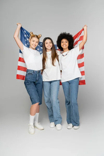 Повна довжина позитивних і багатоетнічних дівчат-підлітків у білих футболках тримає американський прапор і дивиться на камеру на сірому фоні, енергійні друзі-підлітки проводять час, дружба — стокове фото