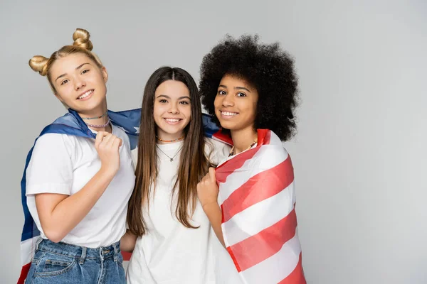 Portrait of positive and multiethnic teenage girlfriends in white t-shirts covering with american flag and looking at camera isolated on grey, energetic teenage friends spending time — Stock Photo