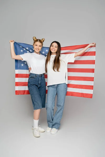 Full length of cheerful blonde and brunette teen girlfriends in white t-shirts holding american flag and standing on grey background, energetic teenage friends spending time, friendship — Stock Photo