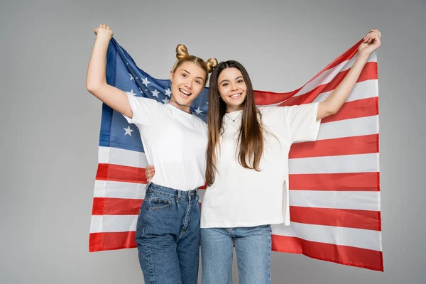 Excited blonde teenage girl in jeans and white t-shirt holding american flag and hugging girlfriend while standing together isolated on grey, energetic teenage friends spending time — Stock Photo