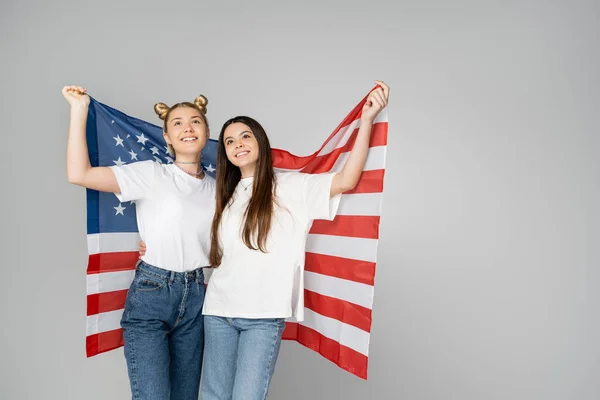 Bruna e bionda adolescenti ragazze in bianco t-shirt e jeans con bandiera americana e guardando in alto mentre in piedi isolato su grigio, energici amici adolescenti trascorrere del tempo, amicizia — Foto stock