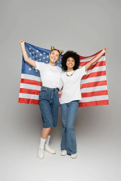 Longitud completa de la chica adolescente rubia sonriente en camiseta blanca que sostiene la bandera americana con amigo afroamericano juntos sobre fondo gris, amigos adolescentes enérgicos pasar tiempo - foto de stock