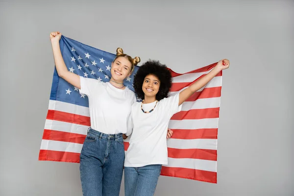 Positive and multiethnic girlfriends in white t-shirts and jeans holding american flag and smiling at camera isolated on grey, energetic teenage friends spending time, friendship and companionship — Stock Photo