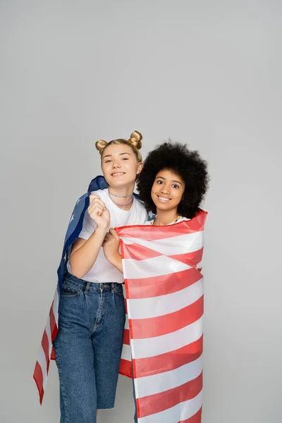 Sorrindo loiras e africanas namoradas adolescentes americanas cobrindo com bandeira americana e olhando para a câmera enquanto estava isolado em cinza, amigos adolescentes enérgicos passar tempo, amizade — Fotografia de Stock