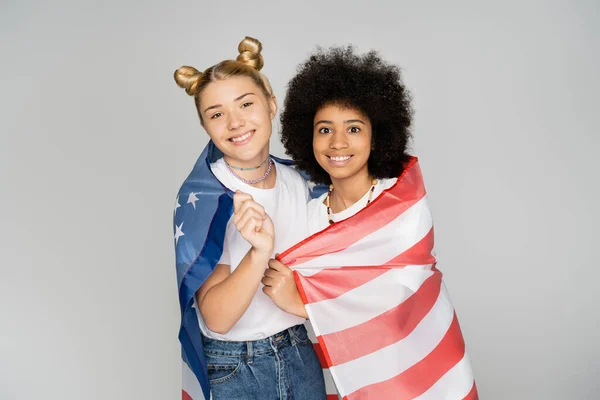 Portrait of smiling blonde and african american teen girlfriends covering with American flag and looking at camera isolated on grey, energetic teenage friends spending time — Stock Photo
