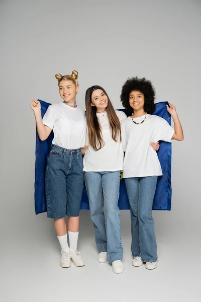 Full length of smiling and multiethnic teenage girlfriends in jeans and white t-shirts holding blue flag and standing on grey background, energetic teenage friends spending time — Stock Photo