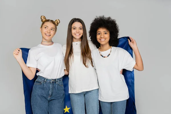 Cheerful and multiethnic teen girlfriends in white t-shirts and jeans holding european flag together and standing isolated on grey, energetic teenage friends spending time, friendship — Stock Photo