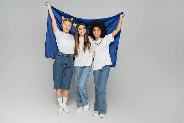 Full length of cheerful interracial teen girlfriends in white t-shirts holding European flag together and standing on grey background, energetic teenage friends spending time — Stock Photo