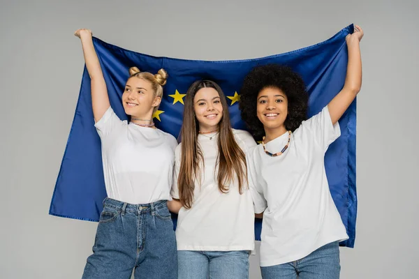 Positive and multiethnic teenage girls in white t-shirts and jeans holding blue european flag while standing together isolated on grey, energetic teenage friends spending time — Stock Photo