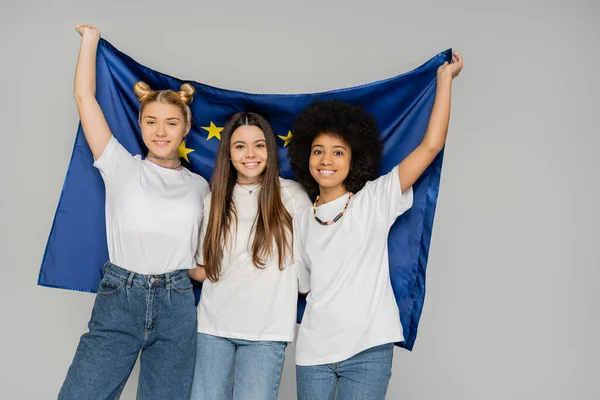 Радостные и многонациональные девочки-подростки в футболках и джинсах, держащие европейский флаг, позируя и стоя изолированно на серых, энергичных друзьях-подростках, проводя время, дружбу и дружбу — стоковое фото