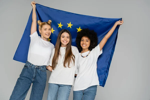 Excited and smiling multiethnic teenagers in white t-shirts and jeans holding blue european flag while posing and standing isolated on grey, energetic teenage friends spending time — Stock Photo