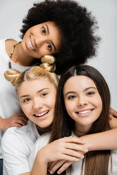 Retrato de namoradas multiétnicas positivas em camisetas brancas abraçando morena amiga adolescente e olhando para a câmera isolada em cinza, amigos adolescentes enérgicos passar tempo, amizade — Fotografia de Stock