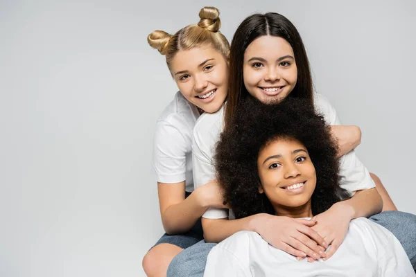Joyful interracial teenage girlfriends in white t-shirts looking at camera while hugging each other and posing isolated on grey, energetic teenage friends spending time, copy space — Stock Photo