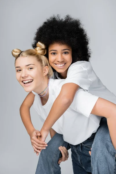 Cheerful african american teenage girl in white t-shirt and jeans piggybacking on blonde friend and having fun isolated on grey, energetic teenage models spending time, friendship and companionship — Stock Photo