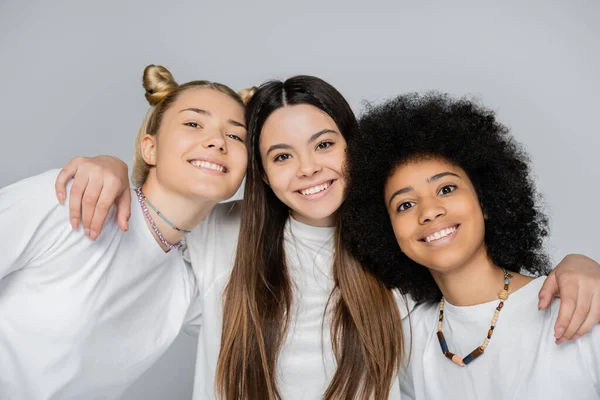 Portrait of positive young brunette teenager hugging interracial girlfriends in white t-shirts and looking at camera isolated on grey, energetic teenage models spending time — Stock Photo