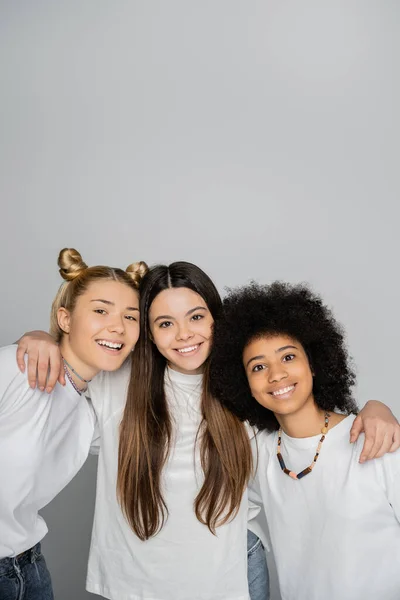 Smiling brunette teen girl in white t-shirt hugging interracial girlfriends and looking at camera together isolated on grey, energetic teenage models spending time, friendship and companionship — Stock Photo