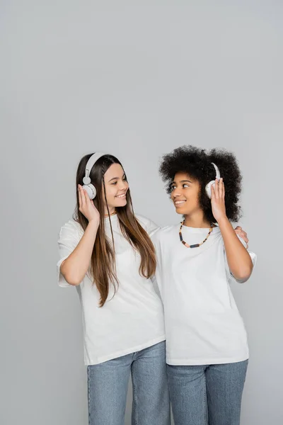 Positive und multiethnische Teenie-Freundinnen in Jeans und weißen T-Shirts, die sich umarmen und mit Kopfhörern Musik hören, isoliert von grauen, energischen Teenie-Models, die Zeit, Freundschaft und Kameradschaft verbringen — Stockfoto