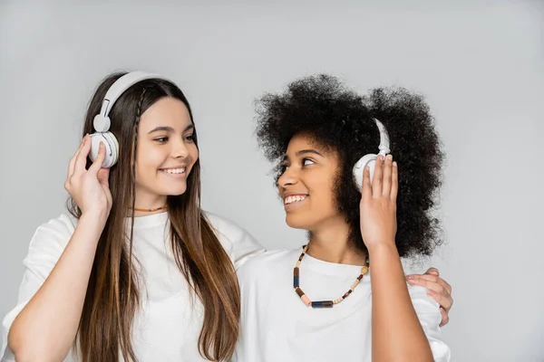 Весела брюнетка дівчина-підліток у білій футболці обіймає афро-американську дівчину з навушниками ізольовані на сірих, енергійних моделях-підлітків, які проводять час, дружбу та товариство — стокове фото