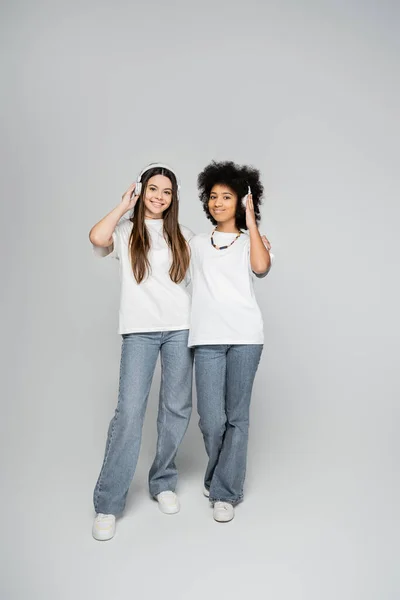 Full length of multiethnic and teenage girlfriends in white t-shirts and jeans listening music in headphones and standing on grey background, energetic teenage models spending time — Stock Photo