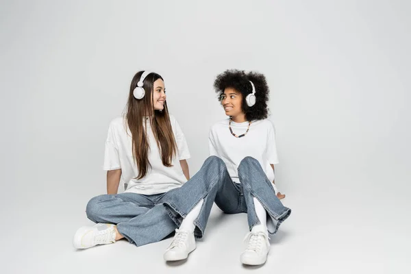 Novias multiétnicas positivas en camisetas blancas, jeans y zapatillas de deporte escuchando música en auriculares y sentadas juntas sobre un fondo gris, adolescentes unidos por el interés común - foto de stock