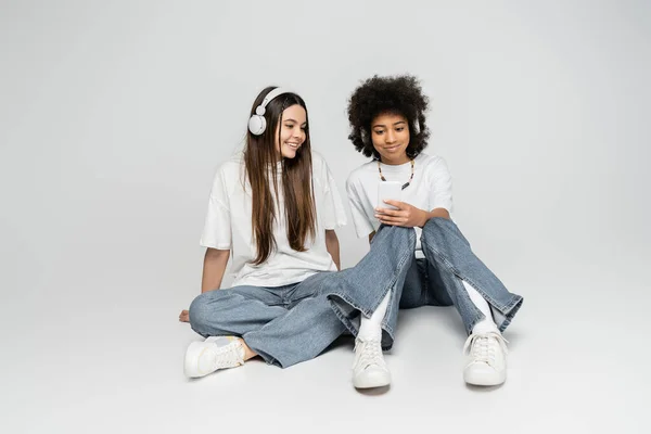 Amigas adolescentes sonrientes y multiétnicas con camisetas blancas y vaqueros usando auriculares y teléfonos inteligentes mientras están sentados en un fondo gris, los adolescentes se unen por el interés común - foto de stock