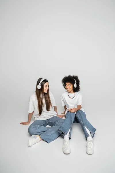 Allegre ragazze adolescenti multietniche in jeans e t-shirt bianche che ascoltano musica in cuffia e usano smartphone su sfondo grigio, adolescenti che legano per interesse comune — Foto stock