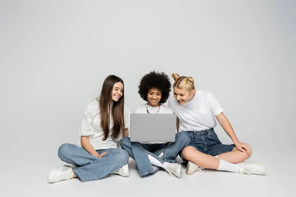 Позитивні багатоетнічні подружки в білих футболках і джинсах, використовуючи ноутбук разом, сидячи на сірому фоні, підлітки зв'язуються над спільними інтересами, дружбою і товариством — стокове фото