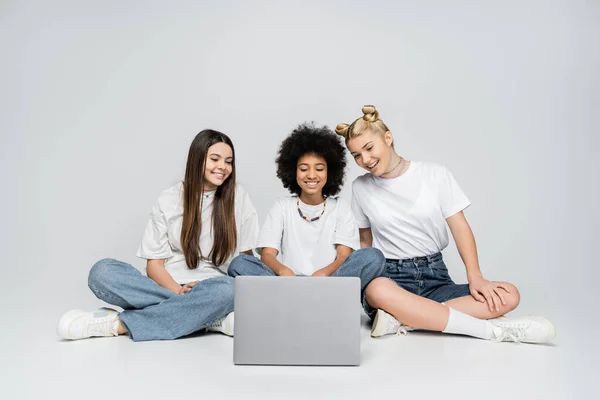 Amigos adolescentes alegres e multiétnicos em camisetas e jeans brancos olhando para o laptop juntos e sentados em fundo cinza, adolescentes se unindo sobre o interesse comum, amizade e companheirismo — Fotografia de Stock