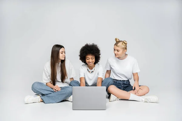 Посмішка підліткової афроамериканської дівчини, яка використовує ноутбук біля дівчат у білих футболках і джинсах, сидячи разом на сірому фоні, підлітки прив'язуються до загального інтересу. — стокове фото